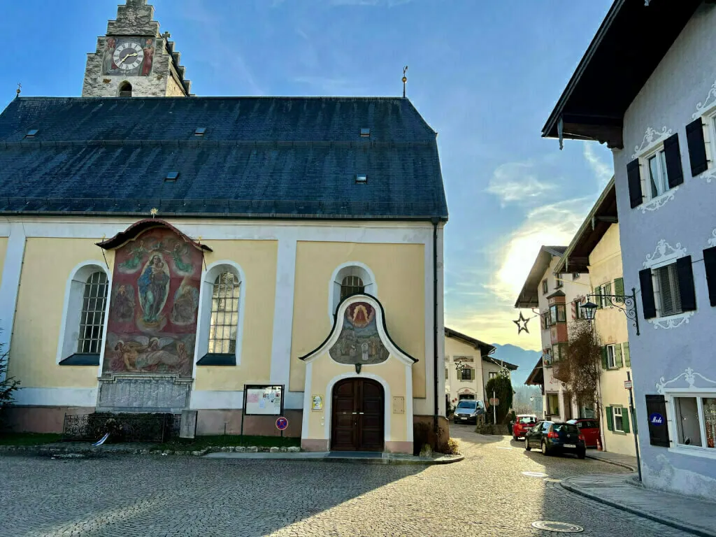 Die Dorfkirche am gepflasterten Platz mit großem religiösem Wandgemälde an der Außenfassade.