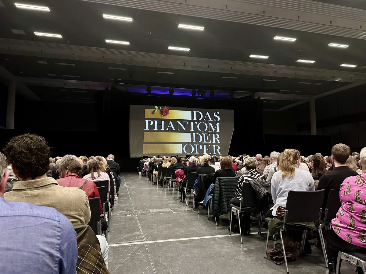 Eine große Menge von Zuschauern sitzt in einem Veranstaltungsraum, der auf eine Bühne ausgerichtet ist. Auf einer Leinwand im Hintergrund steht der Titel "Das Phantom der Oper" in großen Buchstaben und es sind dekorative Elemente sichtbar.