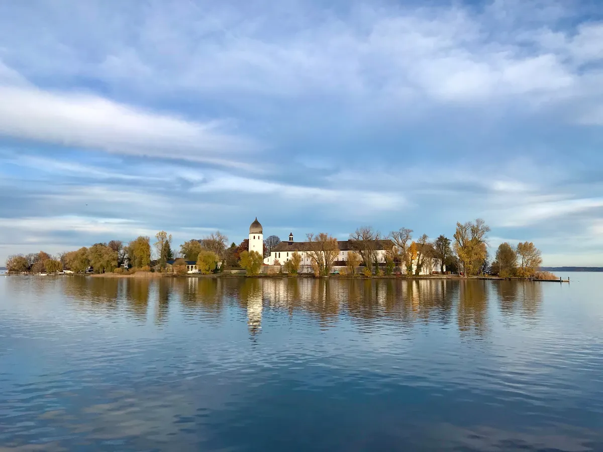 Die malerische herbstliche Fraueninsel spiegelt sich ruhigen Wasser des Chiemsee unter einem bewölkten Himmel.