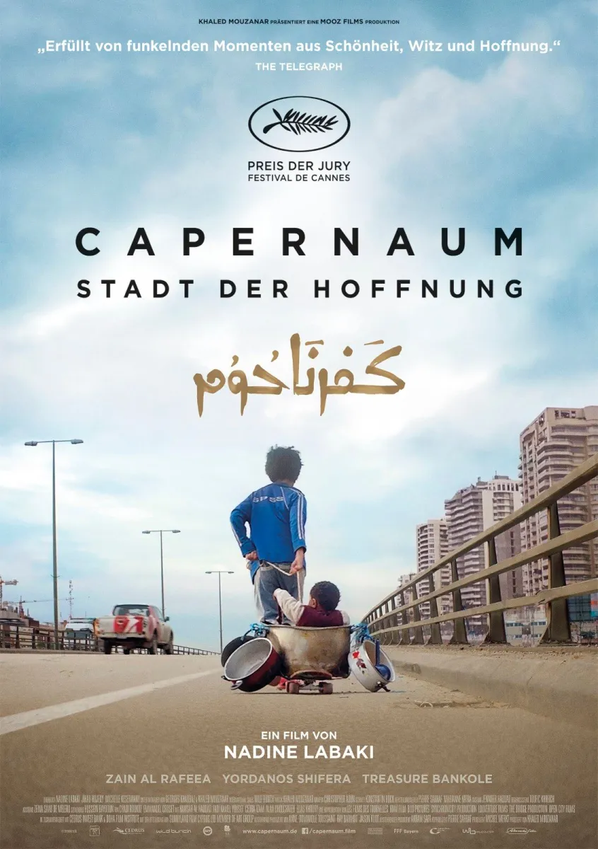 Filmposter für "CAPERNAUM STADT DER HOFFNUNG". Ein Junge steht auf einer erhöhten Brücke und blickt nach unten auf ein kleineres Kind, das in einem Metalltopf sitzt. Sie stehen auf einer leeren Straße, mit einem Auto in der Ferne und hohen Wohnblocks auf der rechten Seite, unter einem hellen, bewölkten Himmel.