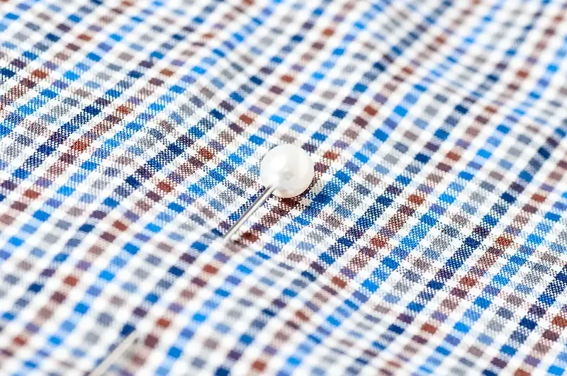 Ein Karomuster mit blauen, braunen und weißen Linien auf einem Stoff. Eine perlweiße Knopf sowie eine Nadel, die durch den Stoff gestochen ist, sind sichtbar.