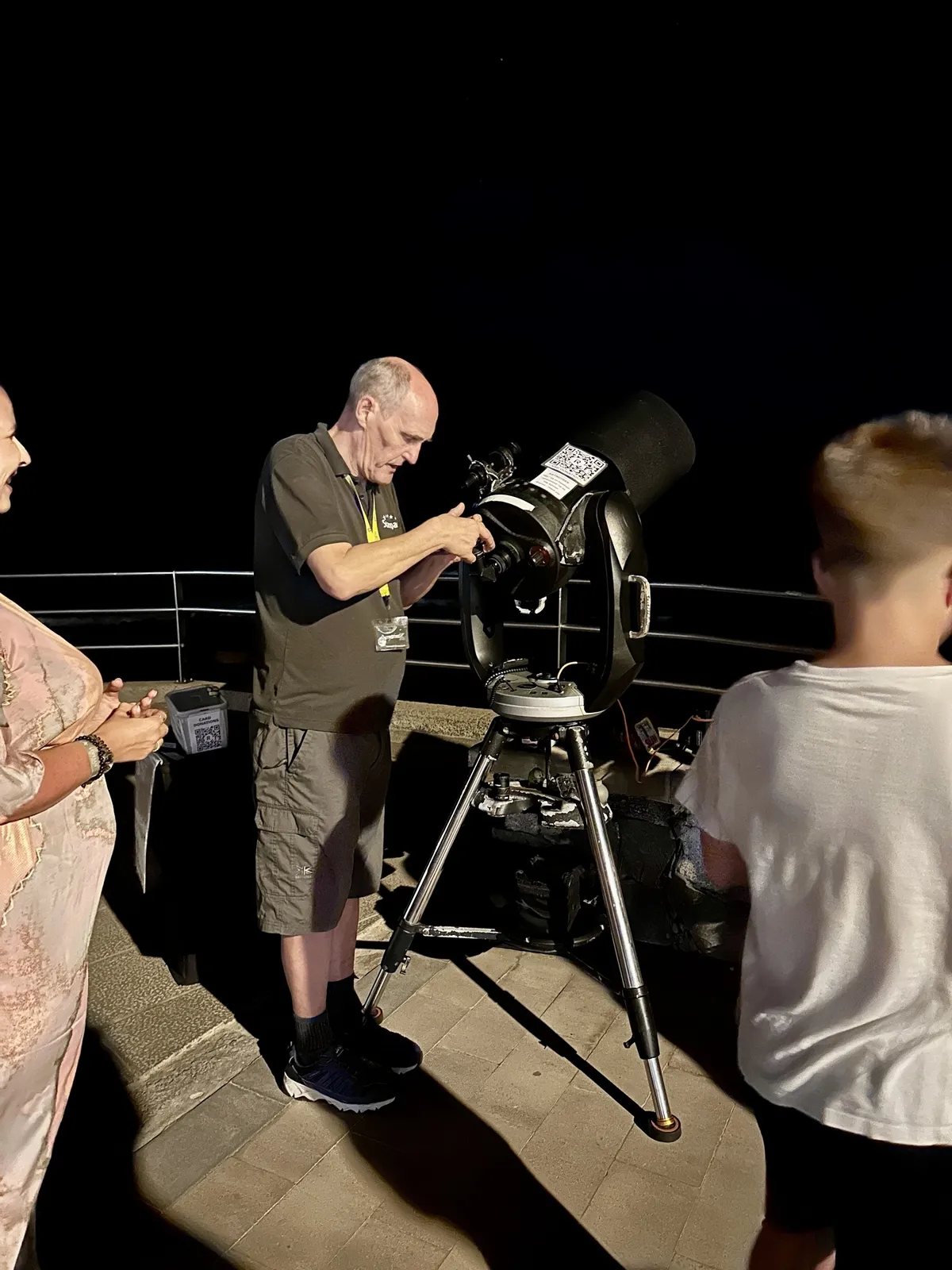 Ein Mann bedient nachts ein Teleskop auf einer Terrasse, während andere Personen zuschauen.