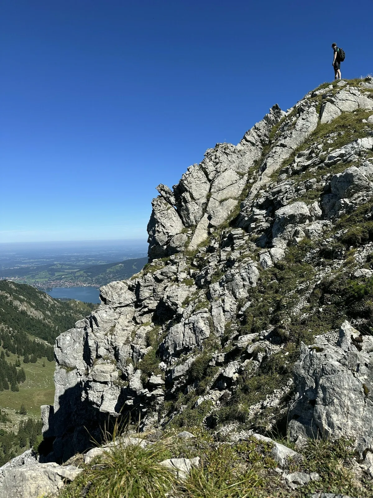Eine steile, felsige Felswand dominiert die Bildmitte. Oben rechts auf dem Gipfel steht eine kleine Person mit Rucksack. Im Hintergrund erstreckt sich eine weite, grüne Tal- und Seenlandschaft unter einem klaren blauen Himmel.