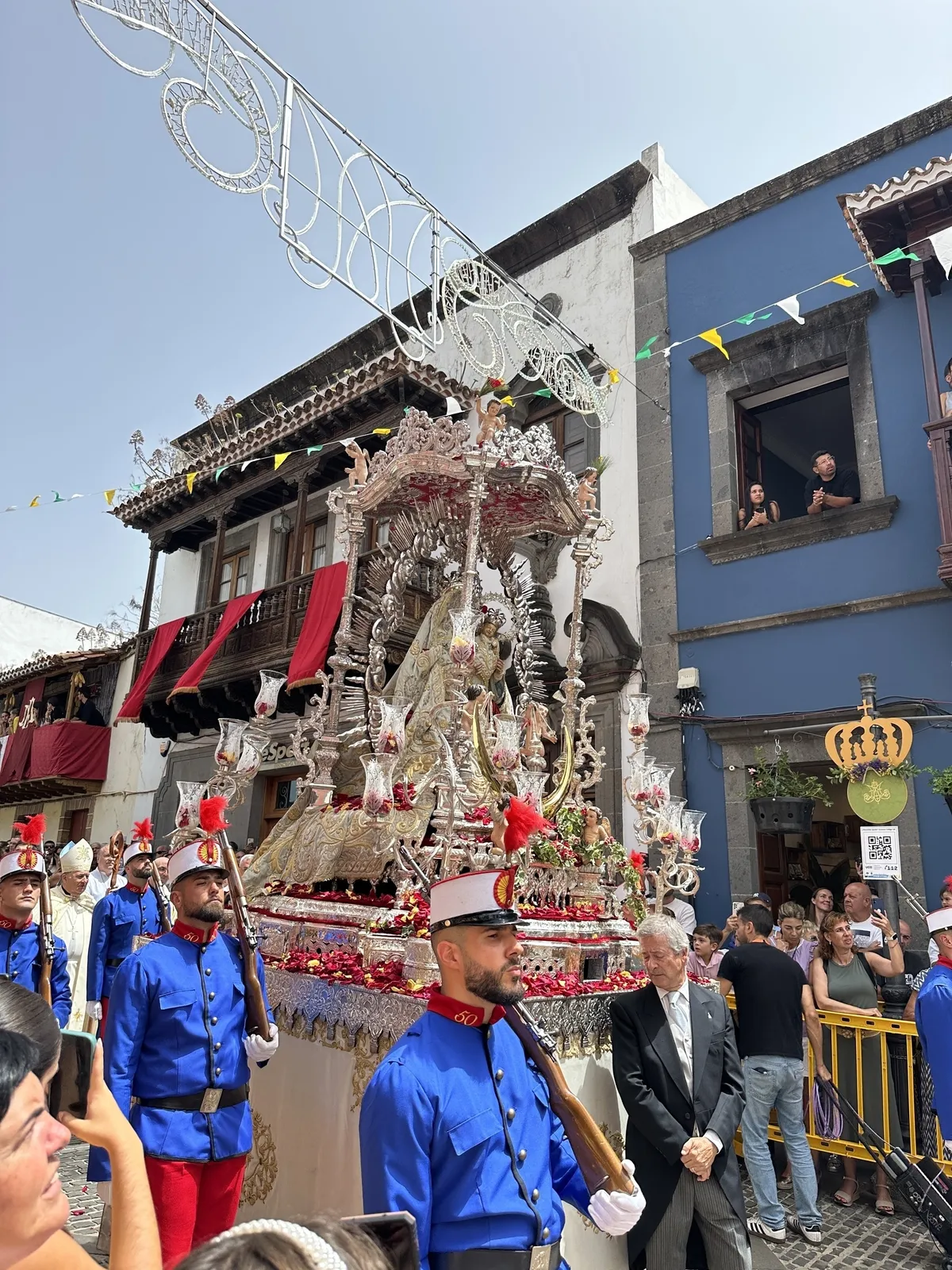 Ein Festzug mit bunt gekleideten Teilnehmern und einem aufwendig geschmückten Wagen zieht durch eine belebte Straße mit Zuschauern, während Menschen aus den Fenstern schauen.