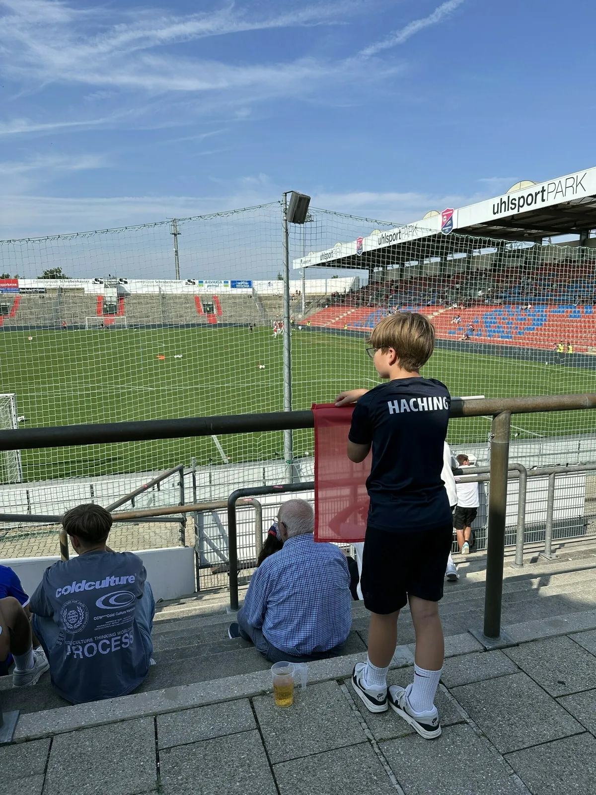 Eine Person in einem dunkelblauen T-Shirt mit der Aufschrift "HACHING" steht in einem Fußballstadion auf einer Stehplatztribüne und blickt auf den Rasen. Neben dieser Person sitzen weitere Zuschauer, darunter eine weitere Person mit einem Bierbecher. Im Hintergrund sind der grüne Rasen, leere rote Sitze und die Überdachung des Stadions unter blauem Himmel zu sehen.