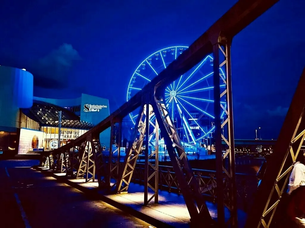 Nachtaufnahme mit einem beleuchteten Riesenrad im Hintergrund, gesehen durch die Stahlkonstruktion einer Brücke am Schokoladenmuseum.