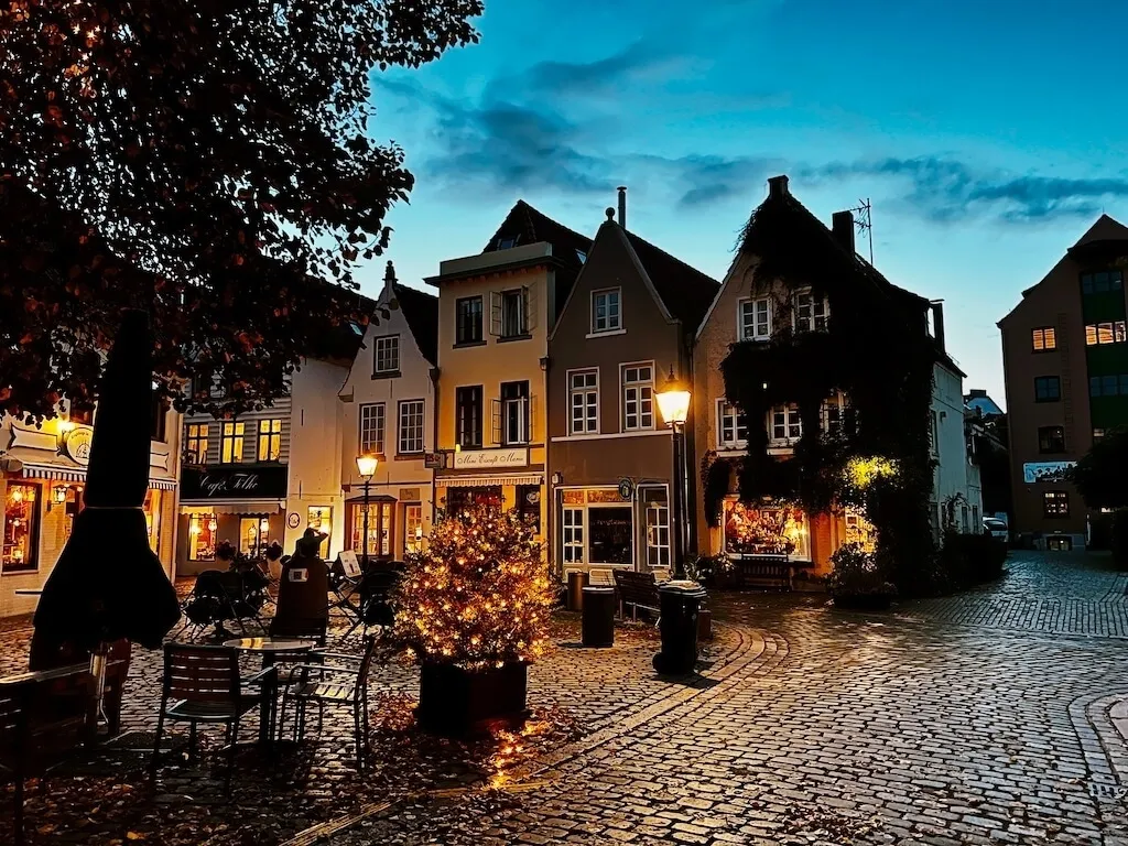 Eine charmante Kopfsteinpflasterstraße in einer abendlichen Altstadt mit beleuchteten Geschäften und dekorativen Lichtern.