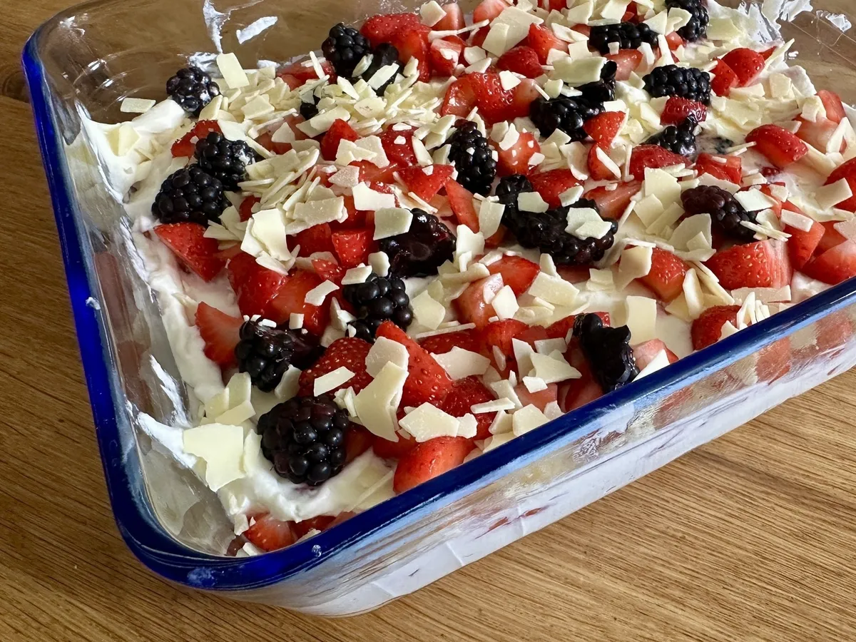 Eine Nachspeise aus Schichten von Früchten, Sahne oder Joghurt und weißer Schokolade in einer Glasform auf einem Holztisch.