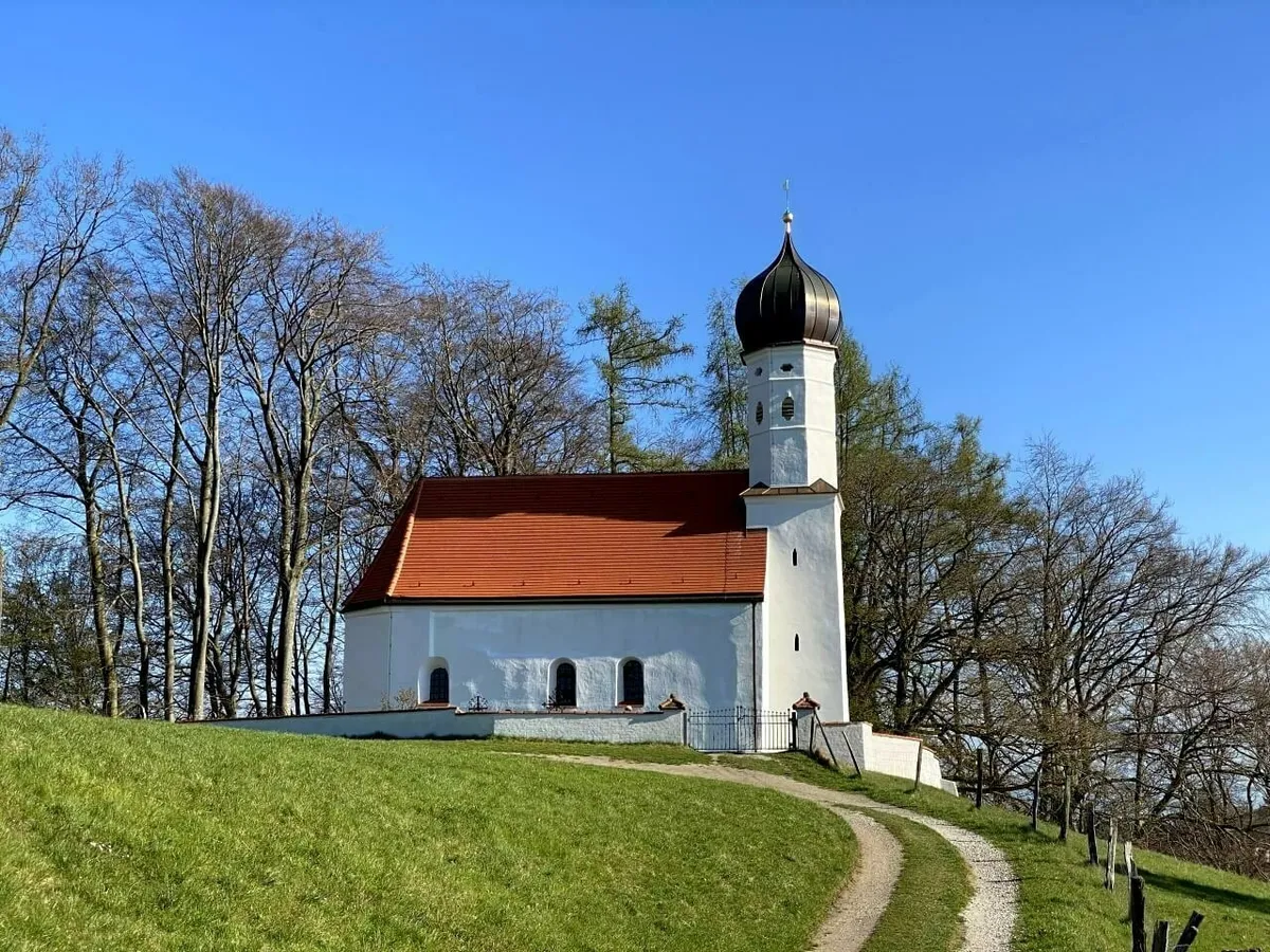 Die weiß getünchte Kirche St. Michael in Ebersberg mit ihrem markanten schwarzen Zwiebelturm, auf einem grünen Hügel gelegen, umgeben von hohen, kahlen Bäumen.