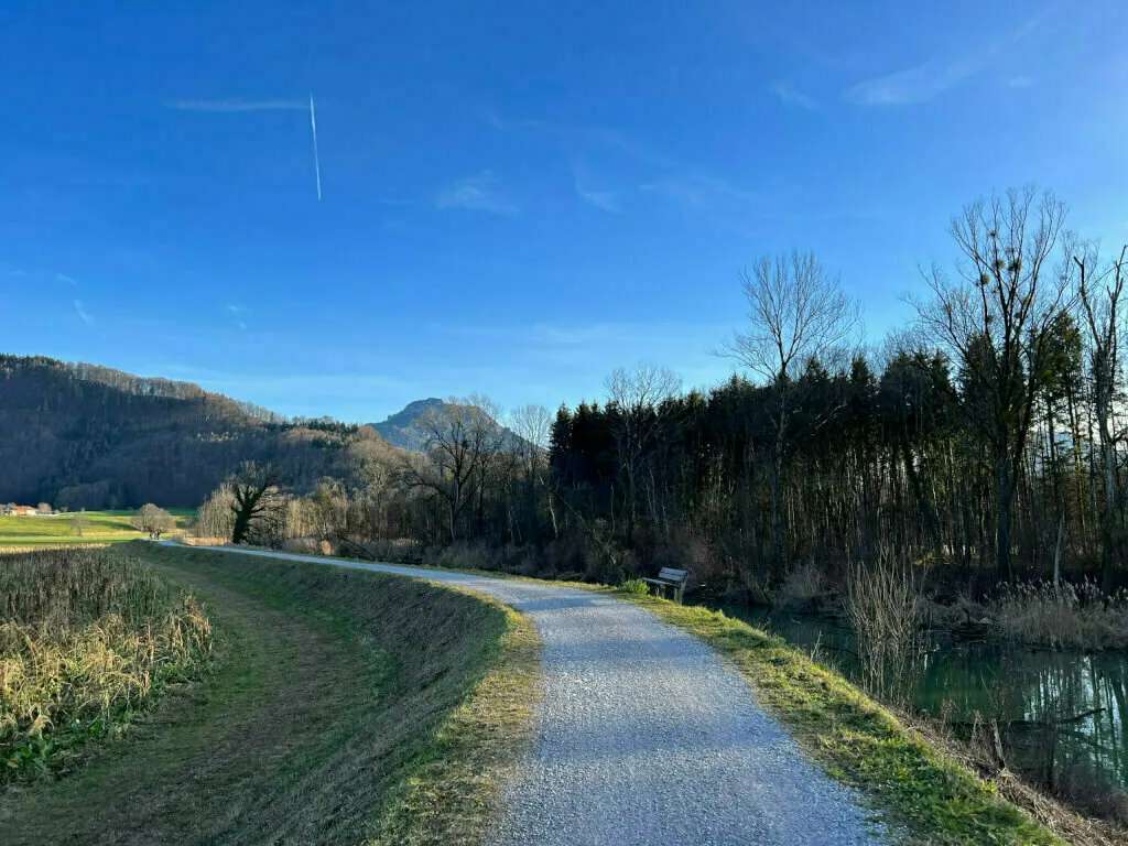 Kiesweg entlang eines Kanals mit Blick auf markante Berggipfel unter blauem Himmel.