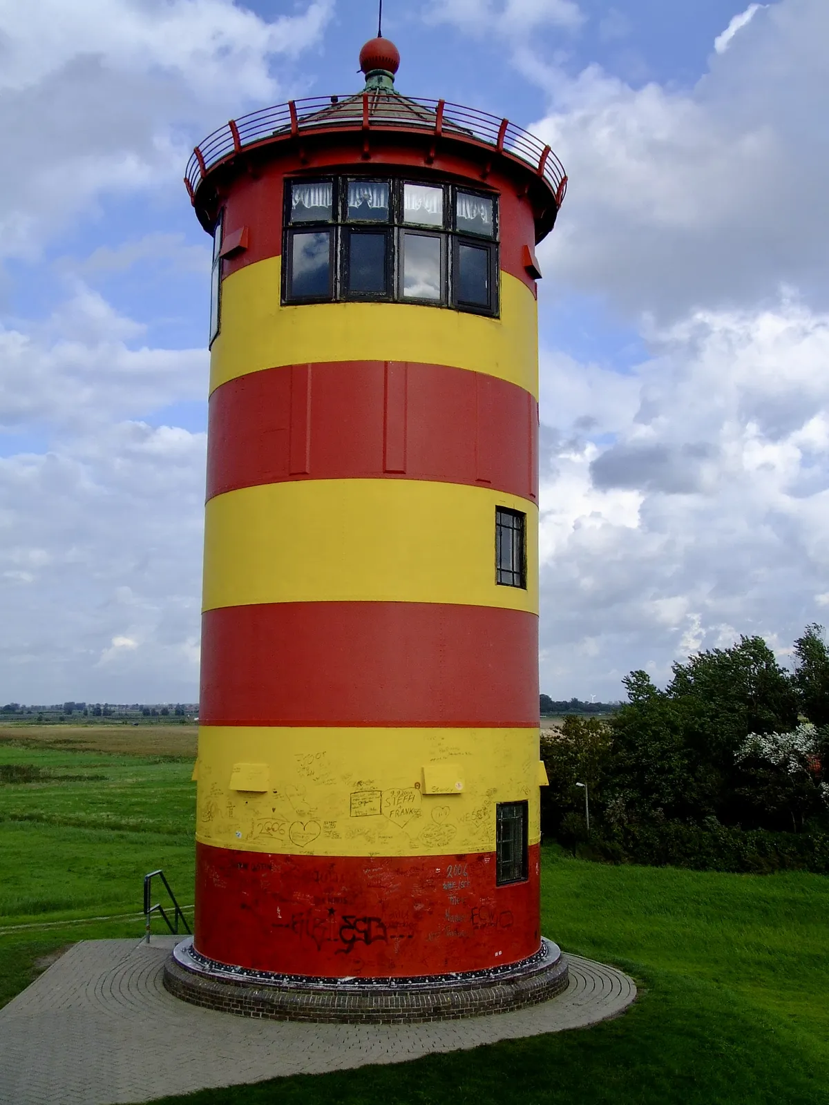 Der auffällig rot-gelb gestreifte Pilsumer Leuchtturm unter einem leicht bewölkten Himmel, umgeben von grünen Wiesen.