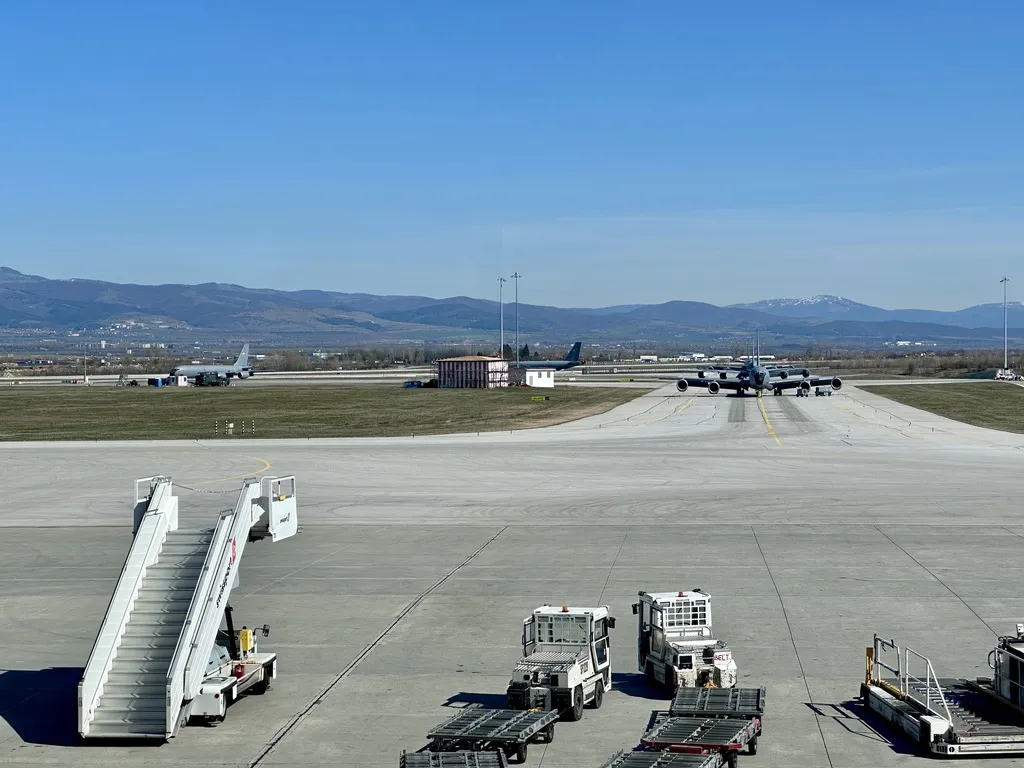 Auf dem Flughafenboden sind Gepäckwagen und eine Treppe zu sehen, während im Hintergrund Flugzeuge auf der Startbahn stehen. Die Landschaft wird von Bergen und klarem Himmel begleitet.