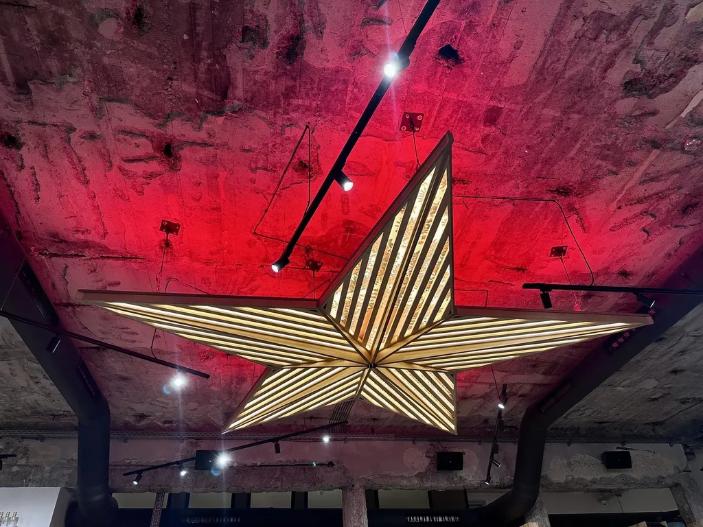 Eine große, hängende Sternleuchte mit geometrischem Design aus Holz und vielen kleinen Lichtern strahlt in einem Raum mit rotem Licht und sichtbaren Betonwänden.