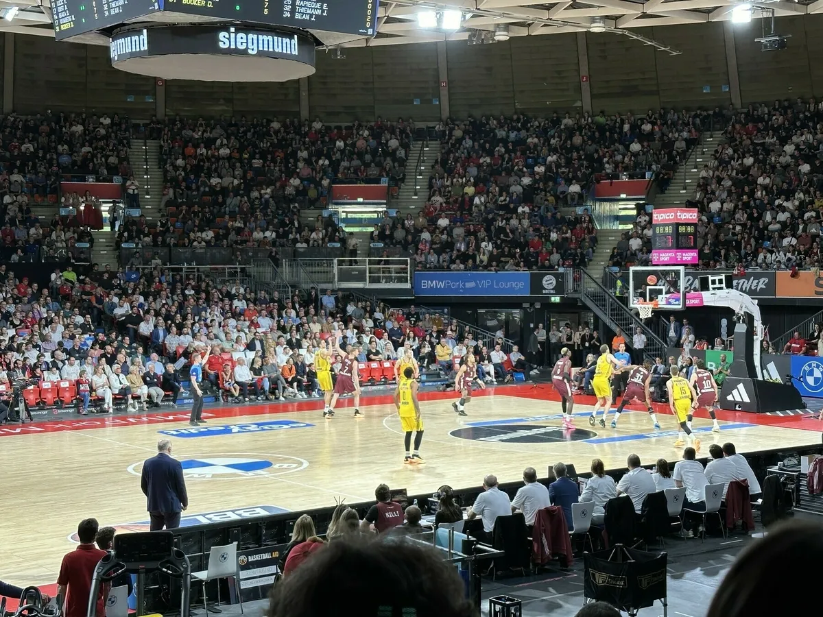 Eine Innenansicht einer gut besuchten Basketball-Arena während eines Spiels. Spieler in gelben und kastanienbraunen Trikots kämpfen um den Ball auf dem Holzplatz.