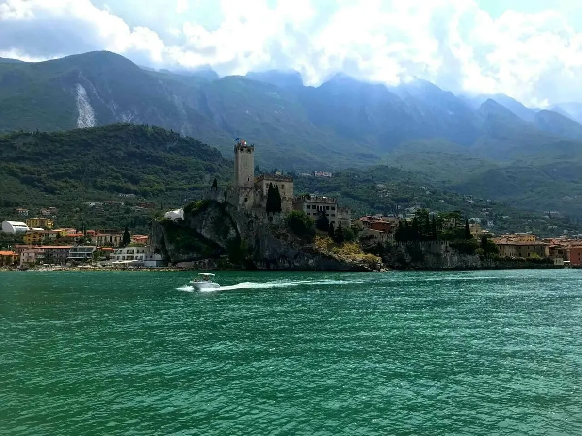Das beeindruckende Scaligerburg (Castello Scaligero) von Malcesine am Gardasee, das auf einem Felsen über dem Wasser thront. Im Vordergrund fährt ein Motorboot vorbei, und dahinter erheben sich majestätische Berge.