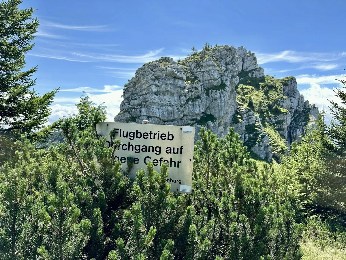 Ein Warnschild mit der Aufschrift „Flugbetrieb, Durchgang auf eigene Gefahr“ ragt aus dichten Latschenkiefern hervor, mit einer großen, zerklüfteten Felsformation im Hintergrund.