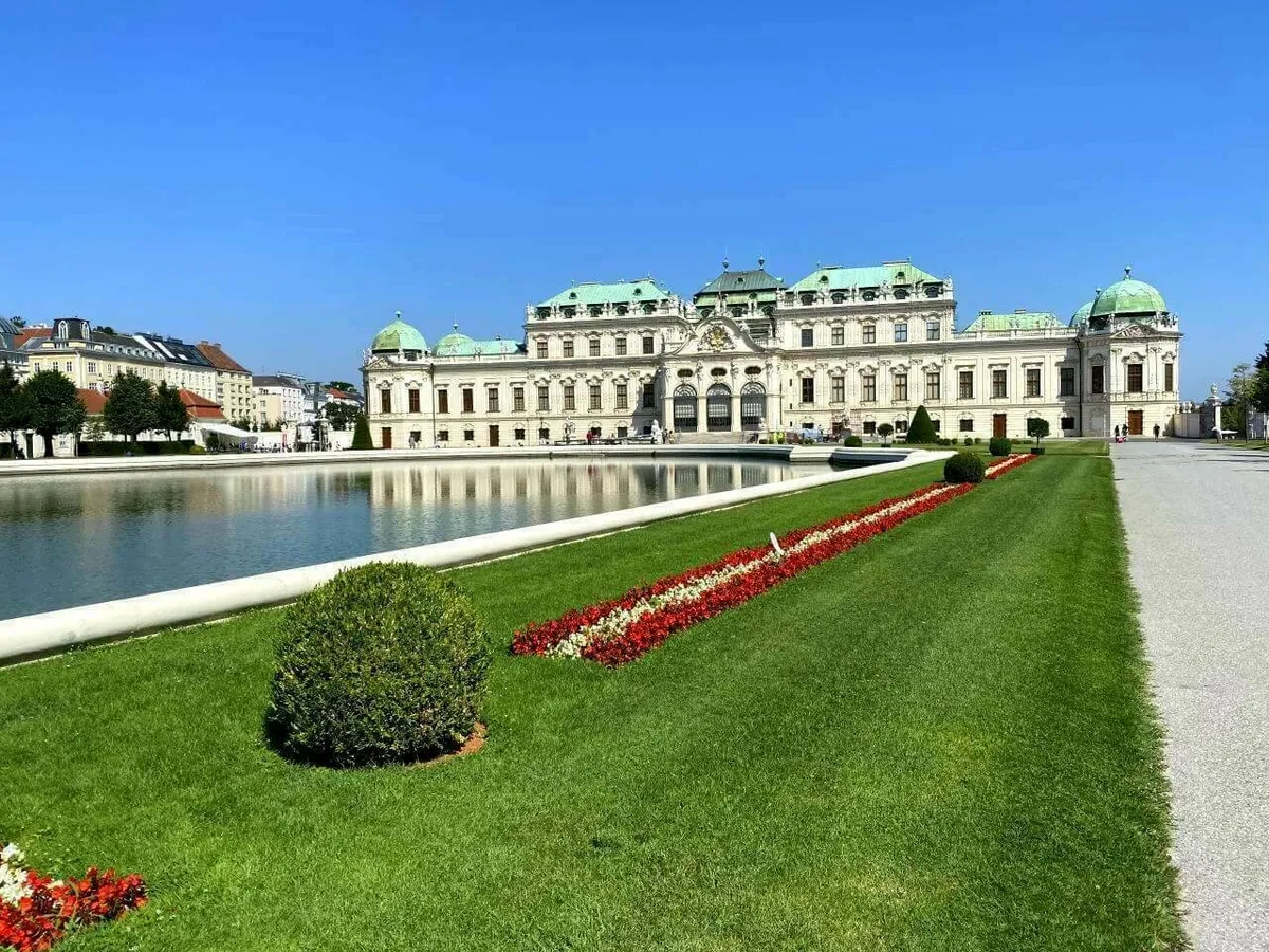Die barocke Fassade des Schloss Belvedere in Wien mit seinen charakteristischen grünen Kuppeln, gespiegelt in einem großen Wasserbecken, davor eine gepflegte Rasenfläche mit Blumen.