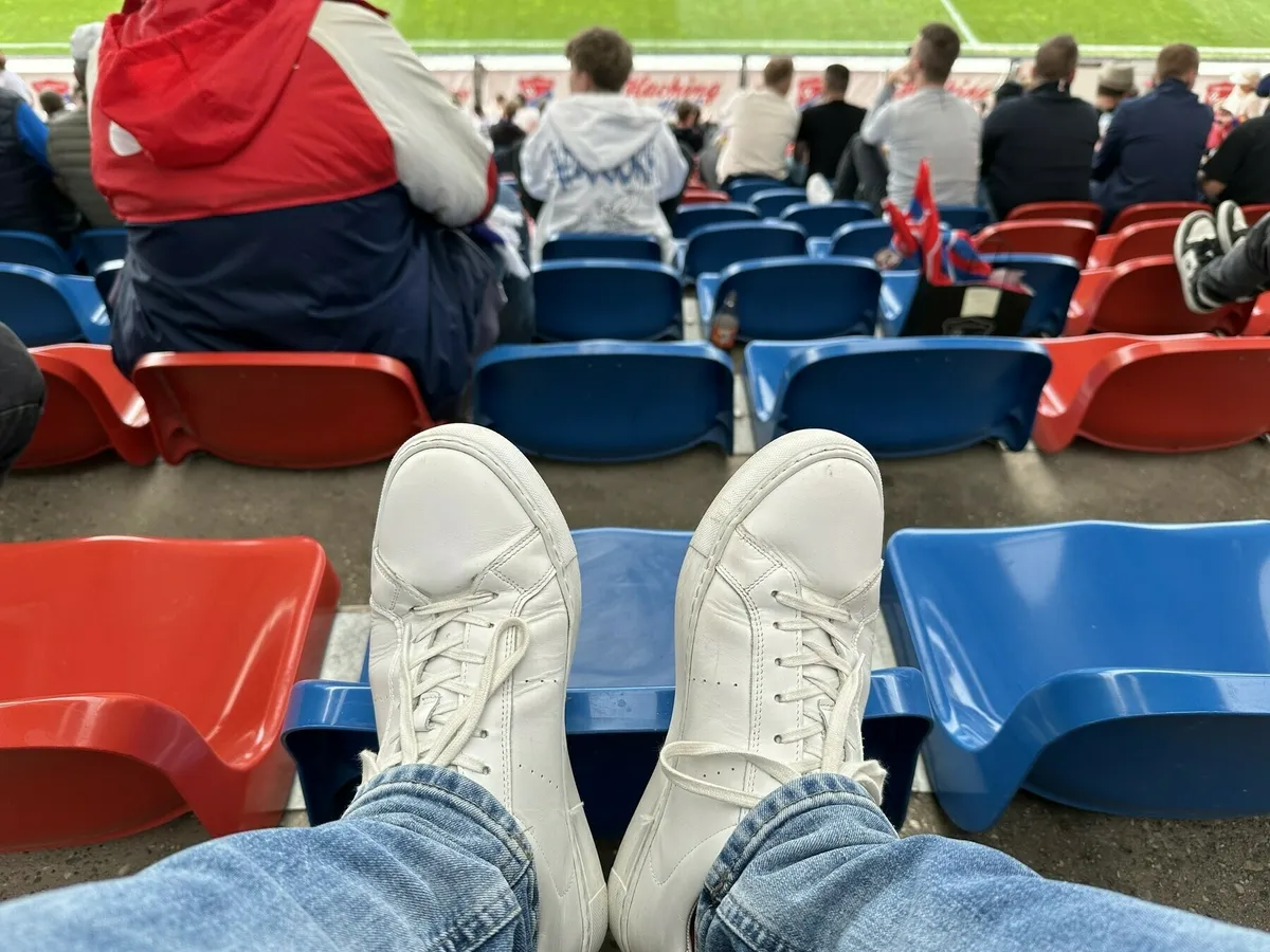 Eine Nahaufnahme der Füße einer Person in weißen Sneakern, die auf den roten und blauen Sitzen eines leeren Stadions ruht, während andere Zuschauer im Hintergrund sitzen.