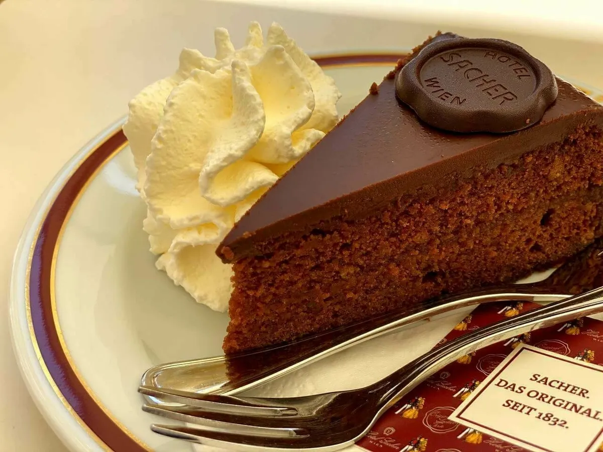 Ein Nahaufnahme-Bild der originalen Sacher Torte vom Hotel Sacher Wien auf einem Teller mit Schlagsahne und dem charakteristischen Schokoladensiegel.
