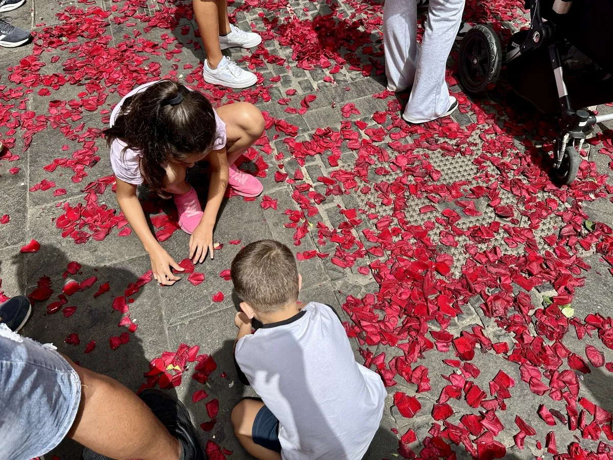 Kinder spielen auf einer mit roten Blütenblättern bedeckten Straße, umgeben von anderen Personen.