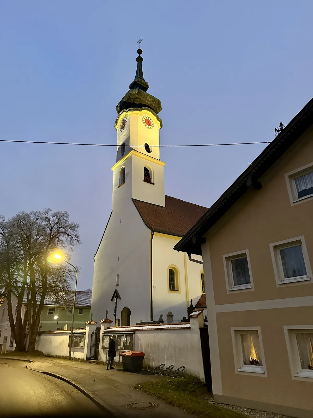 Eine Kirche mit einem hohen, spitzen Turm und einer Uhr in einem ruhigen Abendlicht. Im Vordergrund ist ein Wohnhaus zu sehen, und auf der Straße steht eine Person.