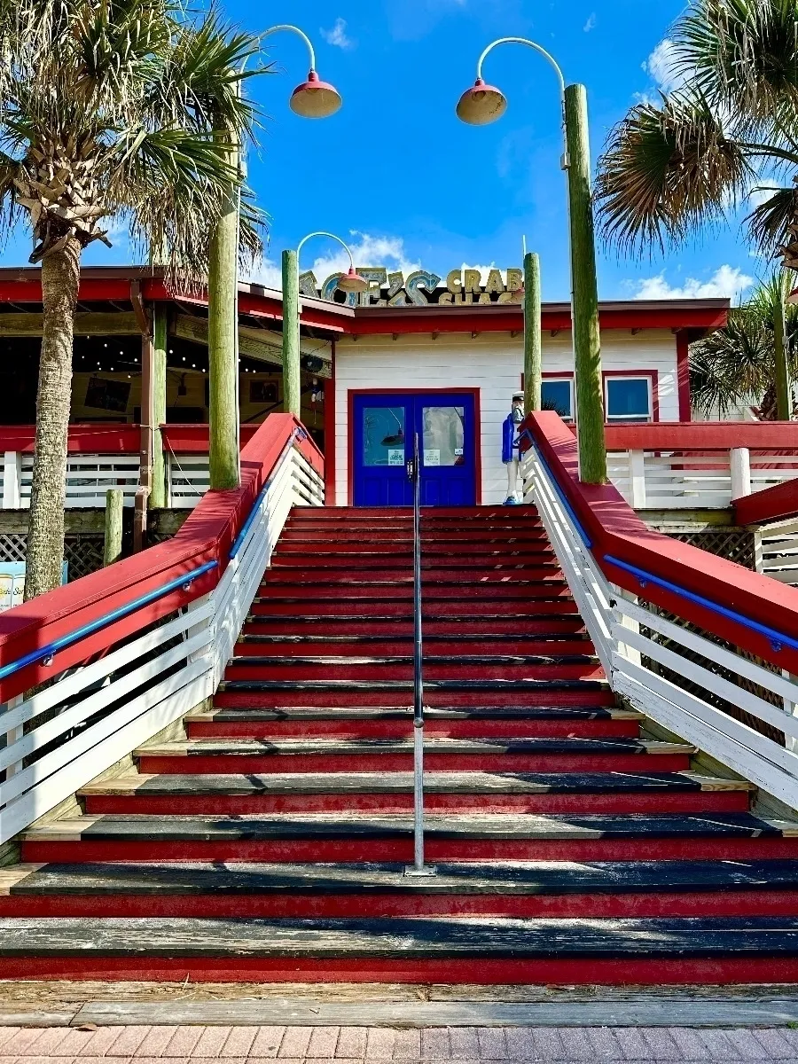 Die Vorderansicht eines Restaurants mit dem Namen "BUCKET OF CRABS" im oberen Bereich, das eine markante, breite Treppe mit roten Stufen und weißen Geländern hat. Links und rechts stehen hohe Palmen.