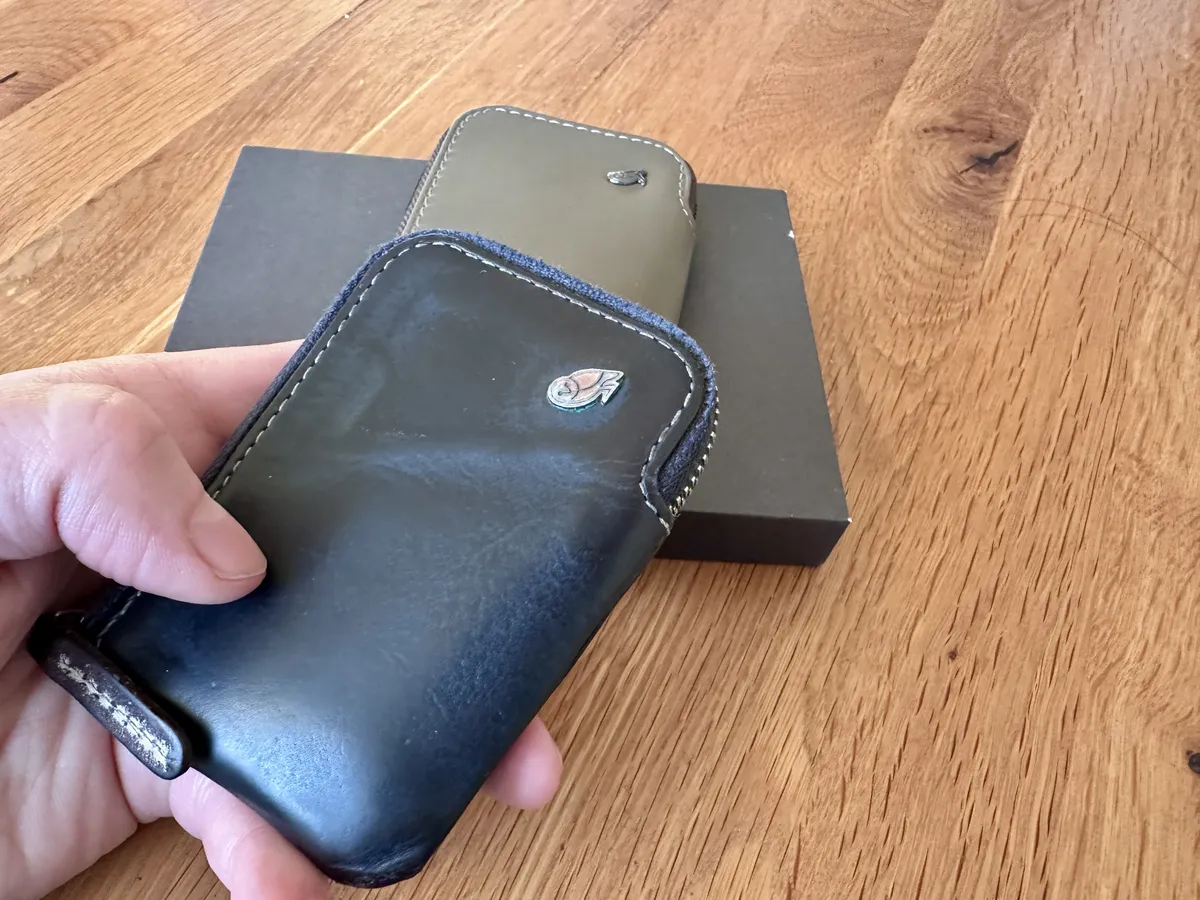 Eine Hand hält ein dunkelblauen Bellroy Card-Pocket während ein grauer Bellroy Card-Pocket dahinter auf einer dunklen Box ruht. Der Holztisch bildet den Hintergrund der Szene.