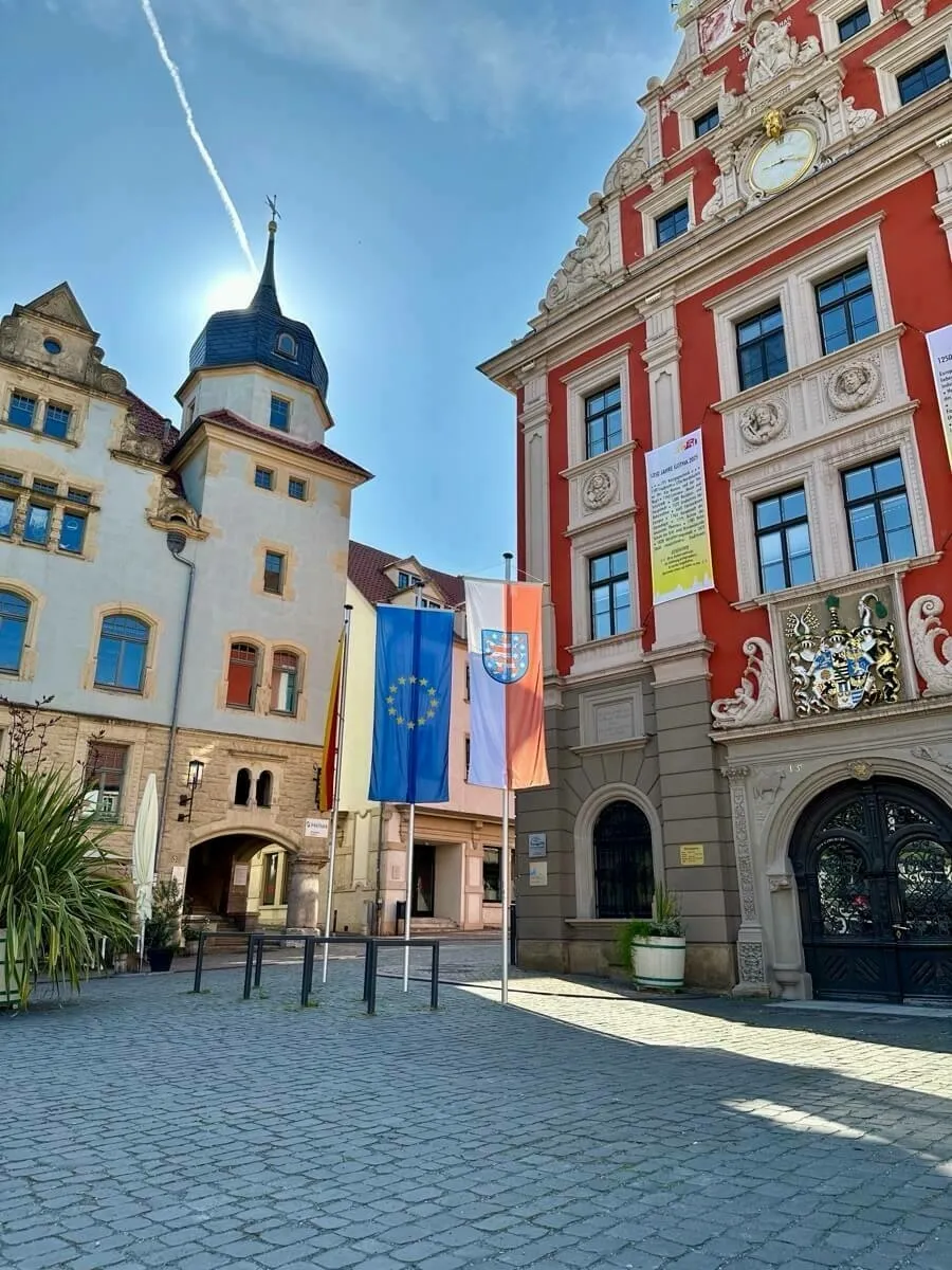 Ein historischer Marktplatz mit beeindruckenden Gebäuden, darunter das historische Rathaus, flankiert von wehenden Fahnen, bei sonnigem Wetter.