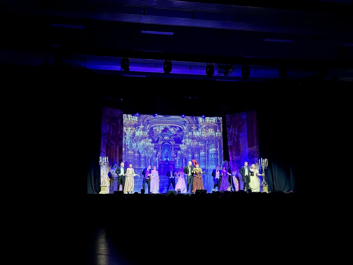 Eine Bühnenaufführung mit mehreren Darstellern in historischen Kostümen, die auf einer eleganten Kulisse stehen. Im Hintergrund sind prachtvolle Kronleuchter und eine opulente Architektur zu sehen, während das Licht die Szenerie in verschiedenen Farben beleuchtet.