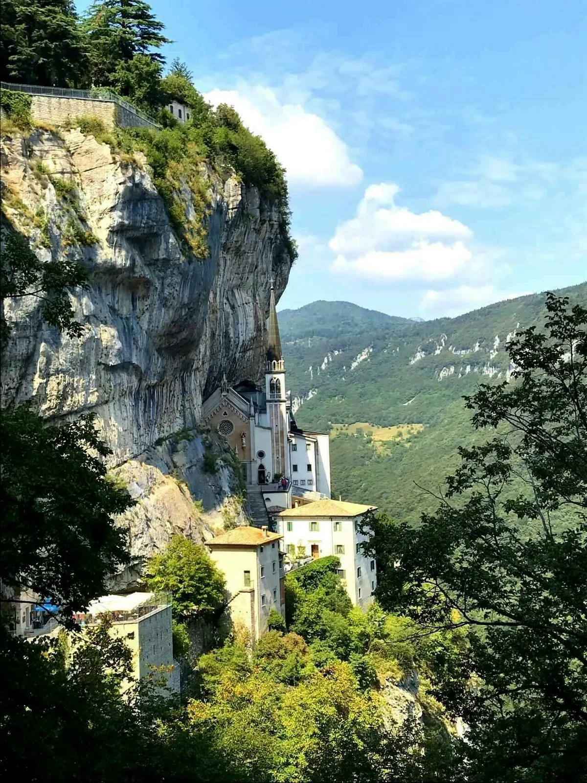 Die Wallfahrtskirche Madonna della Corona, die spektakulär in eine senkrechte Felswand eingebettet ist. Sie ist von grünen Bäumen und Büschen umgeben, und im Hintergrund sind weitere Berge zu sehen.