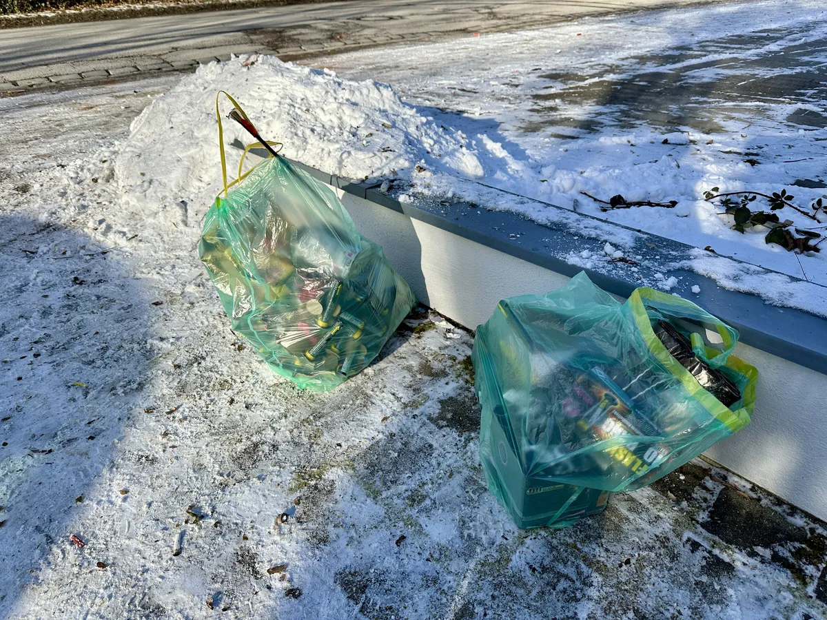 Zwei transparente Müllsäcke vollgestopft mit Silvestermüll stehen in einer verschneiten Einfahrt.