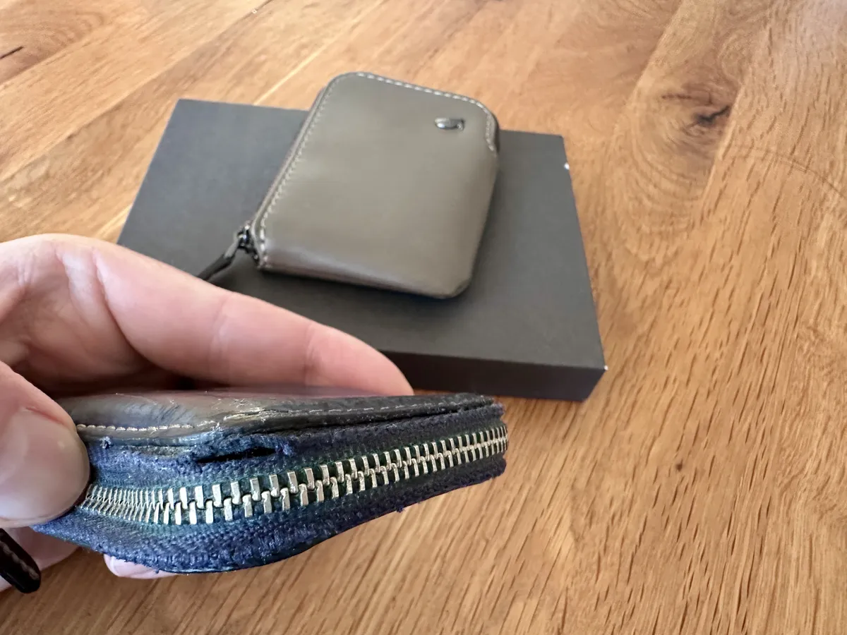 Eine Hand hält eine abgenutzte Geldbörse mit einem sichtbaren Reißverschluss. Im Hintergrund liegt eine neue, graue Geldbörse auf einem schwarzen Karton.