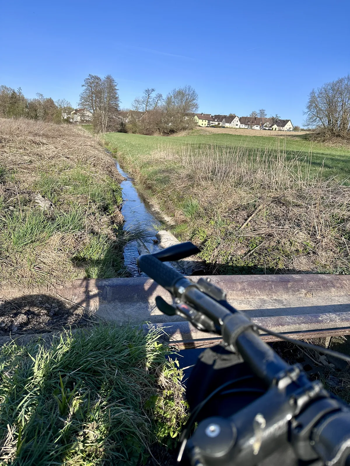 Im Vordergrund ist der Lenker eines Fahrrads zu sehen, während im Hintergrund eine schmale Wasserführung durch eine Wiese verläuft. In der Ferne sind einige Gebäude zwischen Bäumen sichtbar, und der Himmel ist blau mit wenigen Wolken.