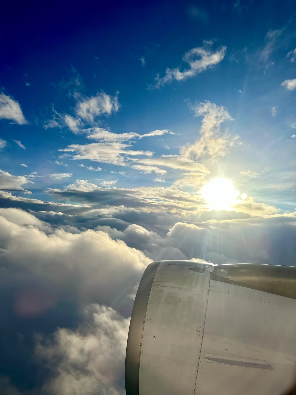 Ein Flugzeugmotor ist im Vordergrund zu sehen, während die Sonne durch eine Schicht aus weißen Wolken und blauem Himmel strahlt.