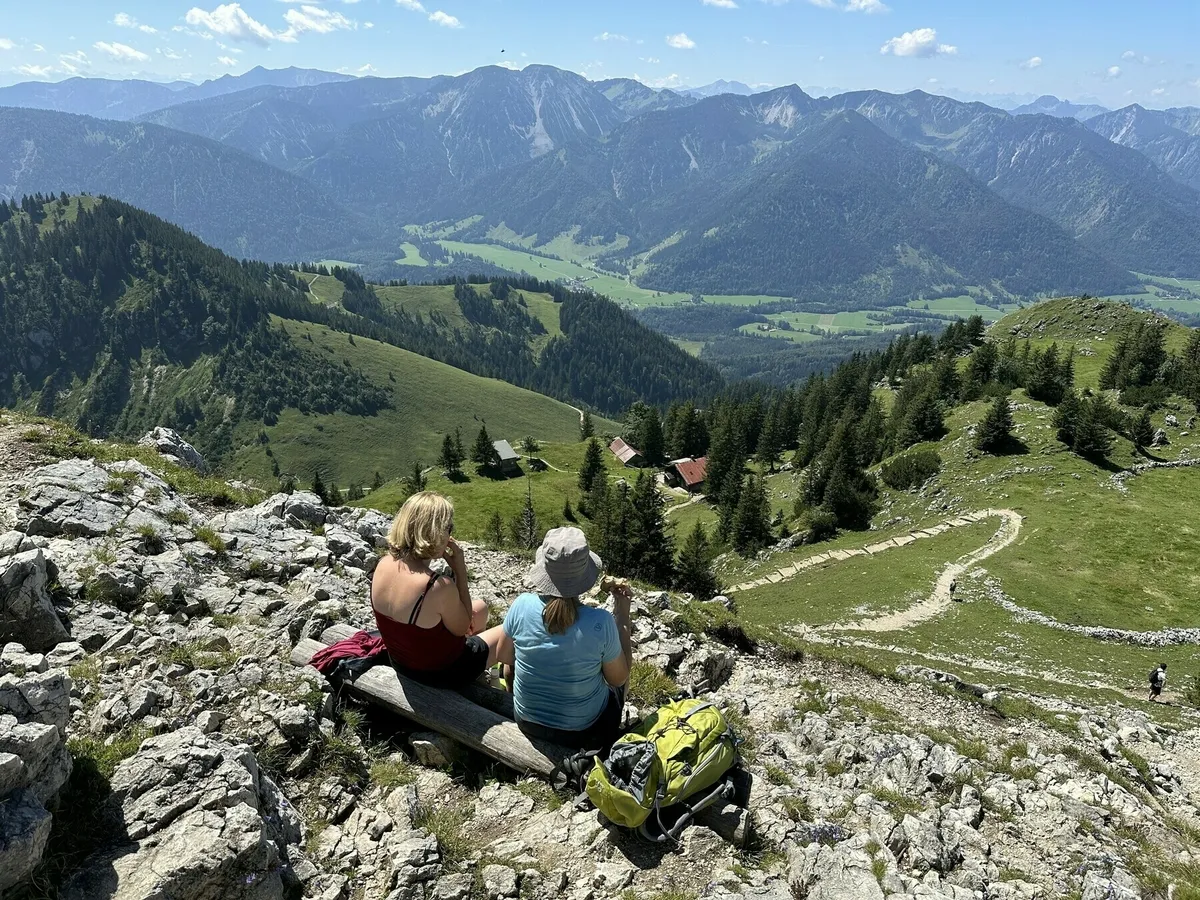 Zwei Wanderinnen sitzen auf einem Felsen am Gipfel und genießen den Blick auf ein grünes Tal und die umliegenden Berge in den Alpen. Im Tal sind eine Almhütte und ein Wanderweg zu sehen.