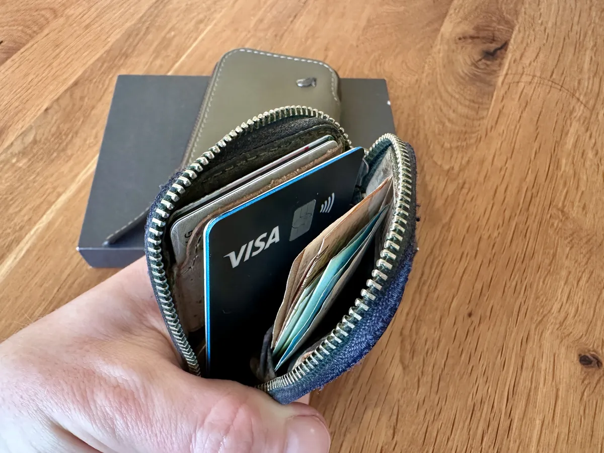Eine Hand hält ein offenes Portemonnaie, das eine Visa-Karte und mehrere Geldscheine sowie weitere Karten sichtbar zeigt. Im Hintergrund ist eine dunkle Box aus Karton zu sehen, auf einem Holztisch platziert.