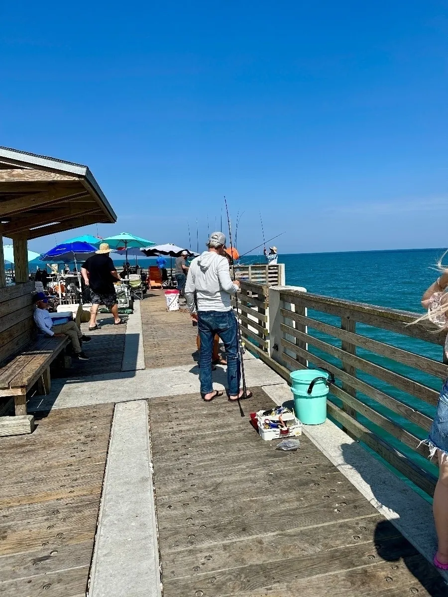 Eine belebte Holzpier an einem sonnigen Tag mit mehreren Leuten beim Angeln. Im Vordergrund steht ein Mann in Jeans und einem grauen Kapuzenpullover. Im Hintergrund sieht man Sonnenschirme und die türkisfarbene See unter einem klaren blauen Himmel.