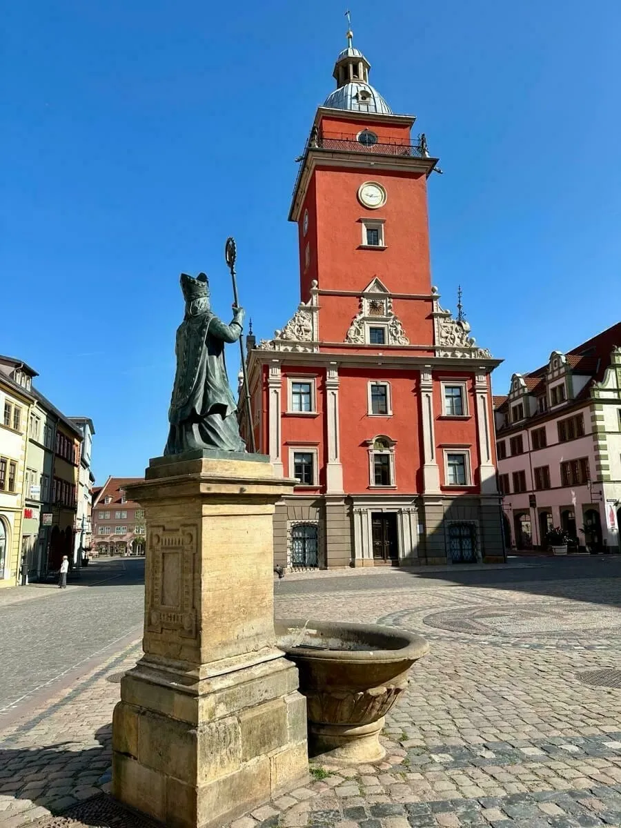 Eine Statue an einem kleinen Springbrunnen steht vor dem historischen Rathaus von Gotha mit Uhrturm in einer gepflasterten Platzlandschaft.