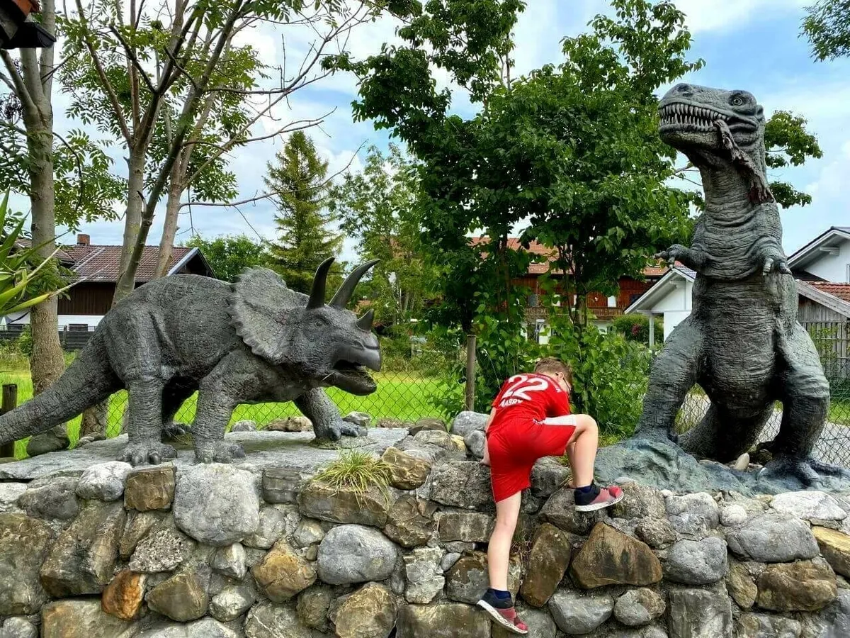 Ein Junge klettert auf einer Felsmauer zwischen zwei großen, grauen Dinosaurier-Statuen, einem Triceratops und ein T-Rex, im Freien.