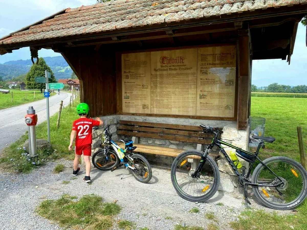 Ein Junge mit Fahrrad steht neben einem Unterstand mit einer Infotafel und zwei abgestellten Fahrrädern auf einem Feldweg.