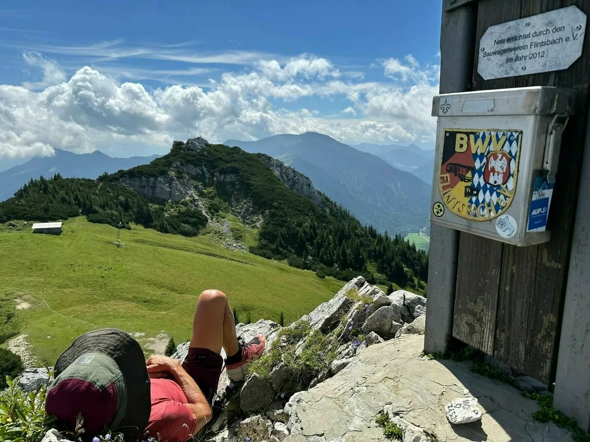 Eine Person liegt auf einem felsigen, grasbewachsenen Gipfel und blickt über eine weite Berglandschaft mit grünen Wiesen und bewaldeten Hügeln. Rechts an einem Holzpfosten ist ein metallener Kasten mit dem Wappen des BWV (Bergwanderverein) Flintsbach e.V. angebracht, darunter eine Plakette, die besagt, dass er 2012 neu errichtet wurde.