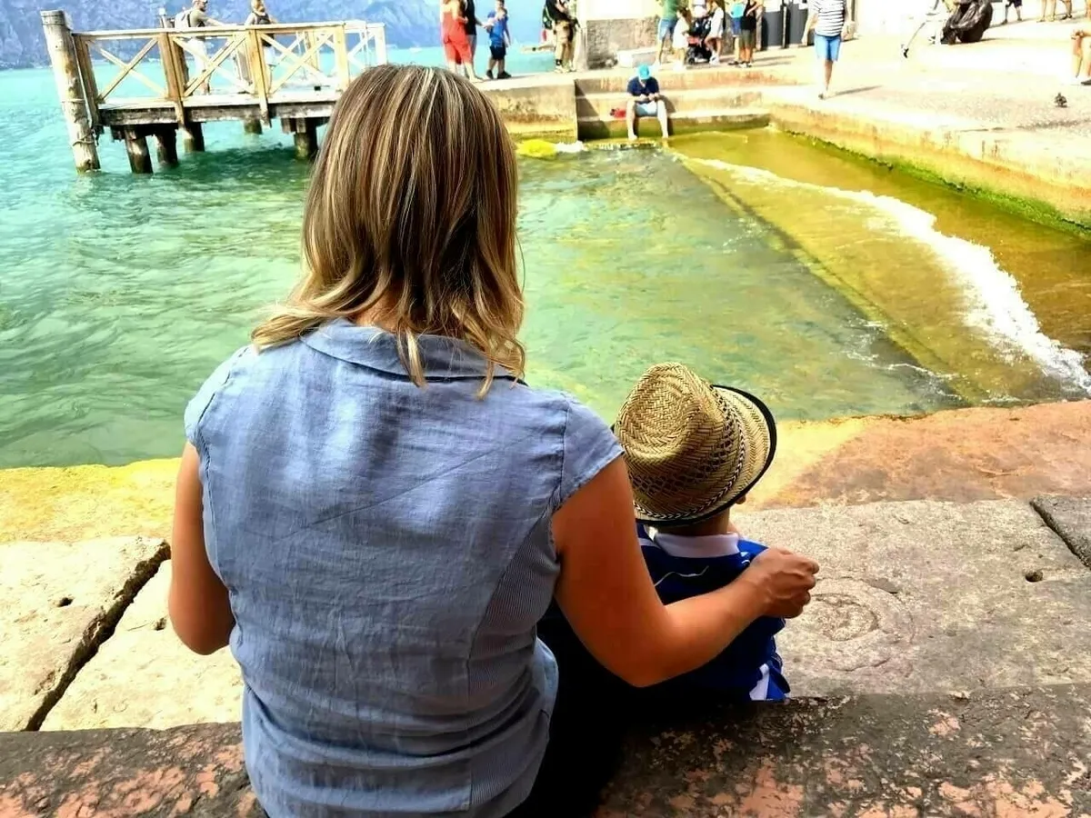 Eine Frau sitzt mit einem Kind, das einen Strohhut trägt, auf einer Steinplattform am Wasser.