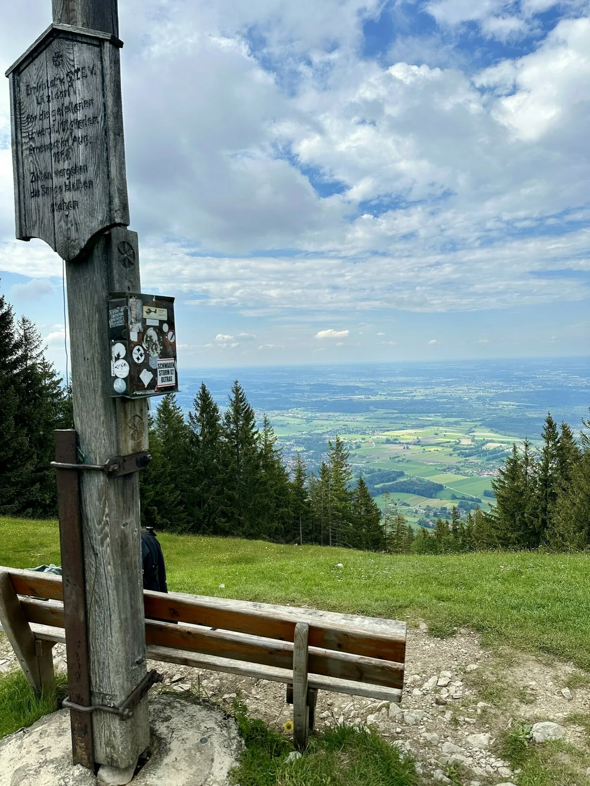Ein rustikaler Holzbalken mit einer geschnitzten Tafel und Aufklebern, vor dem eine Holzbank steht, auf einem Berggipfel. Die Szene bietet einen weiten Ausblick über das flache Land und das Voralpengebiet.