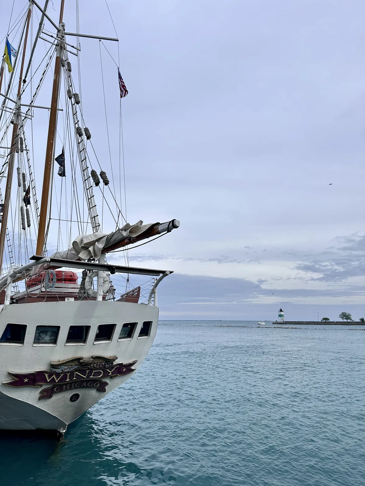 Eine Segelschiff mit mehreren Masten liegt nahe am Ufer eines ruhigen Gewässers. Im Hintergrund ist ein kleiner Leuchtturm und bewölkter Himmel zu sehen.