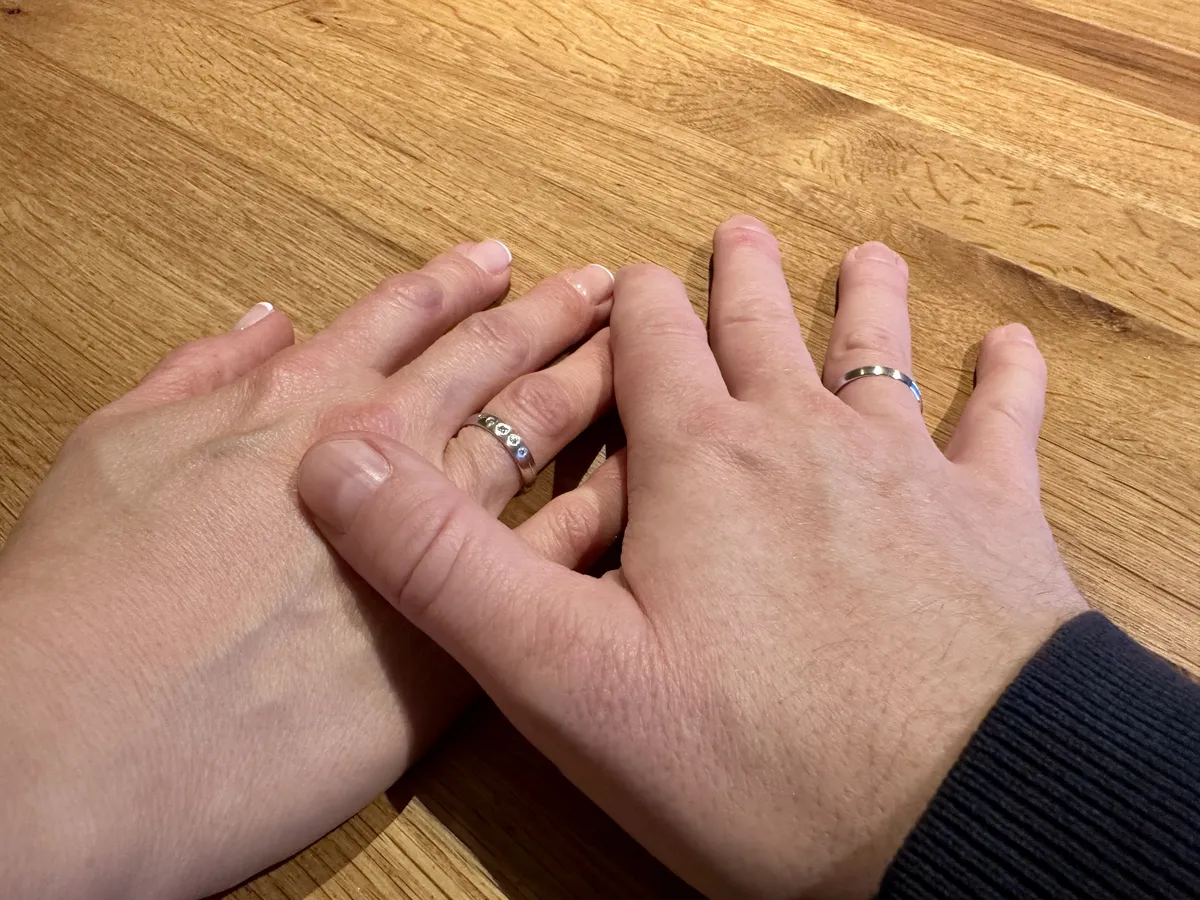 Zwei Hände liegen auf einem Holztisch, wobei die linke Hand einer Person einen schmalen Ring trägt. Die rechte Hand gehört einer anderen Person und zeigt einen Ring sowie einen leichten Unterschied in der Hautfarbe.