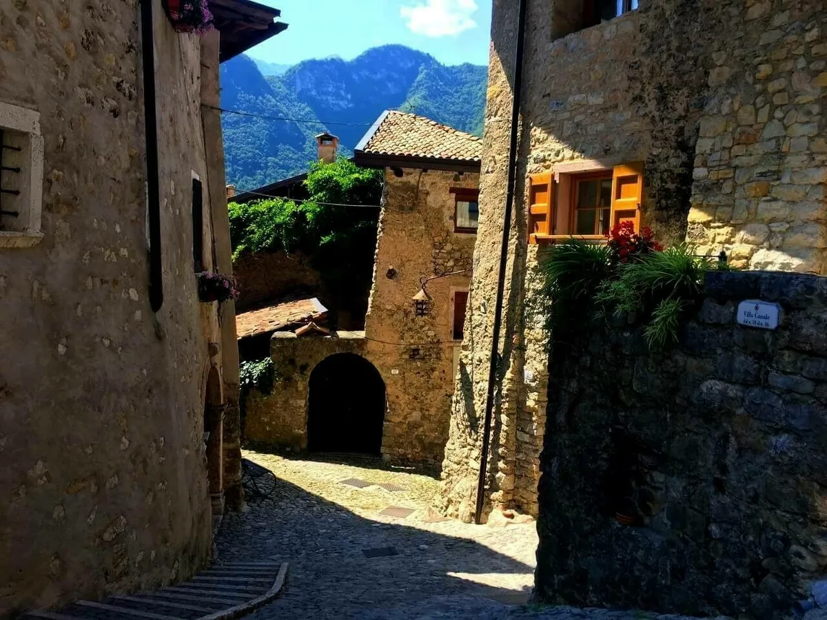 Eine schmale, gepflasterte Gasse im Künstlerdorf Canale di Tenno. Die Häuser bestehen aus Naturstein, und im Hintergrund sind hohe, bewaldete Berge unter blauem Himmel zu sehen.
