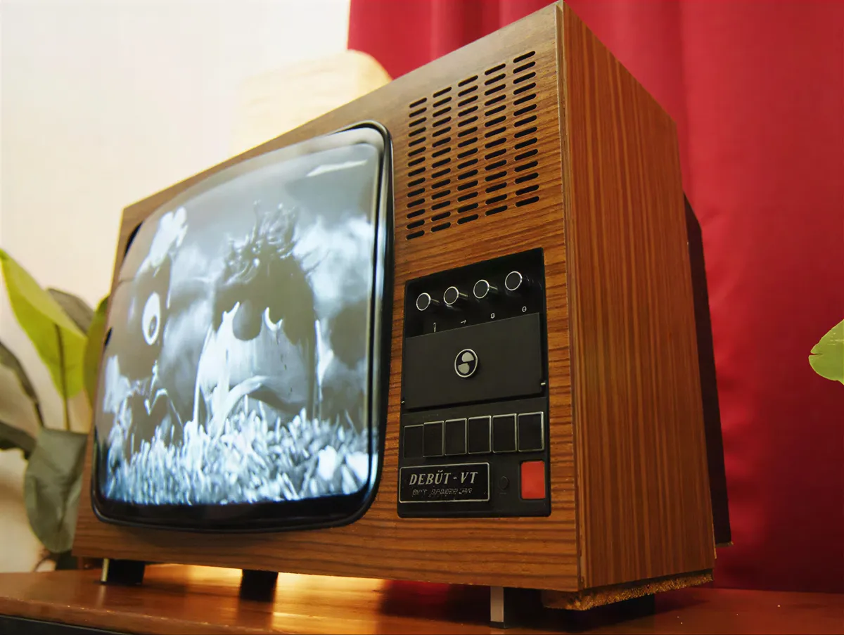 Ein Schwarz-Weiß-Fernseher aus der DDR mit Holzgehäuse, der ein schwarz-weißes Kinderprogramm zeigt. Im Hintergrund sind dekorative Pflanzen und ein roter Vorhang sichtbar.