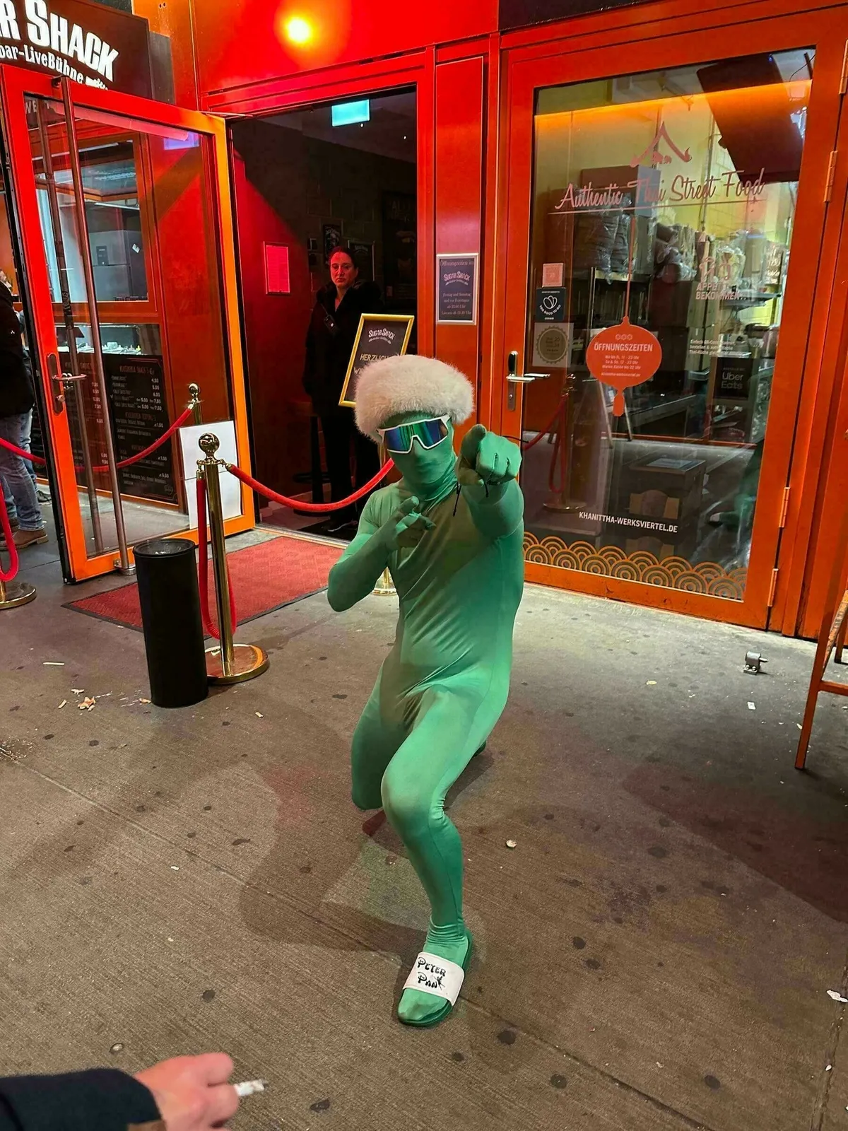Ein Mann in einem hautengen, hellgrünen Ganzkörperanzug, mit weißer Flauschperücke und futuristischer Sonnenbrille, kniet posierend vor dem Eingang eines Restaurants/einer Bar mit roter und oranger Beleuchtung. Er trägt weiße Sandalen mit der Aufschrift "Petsi Pun".