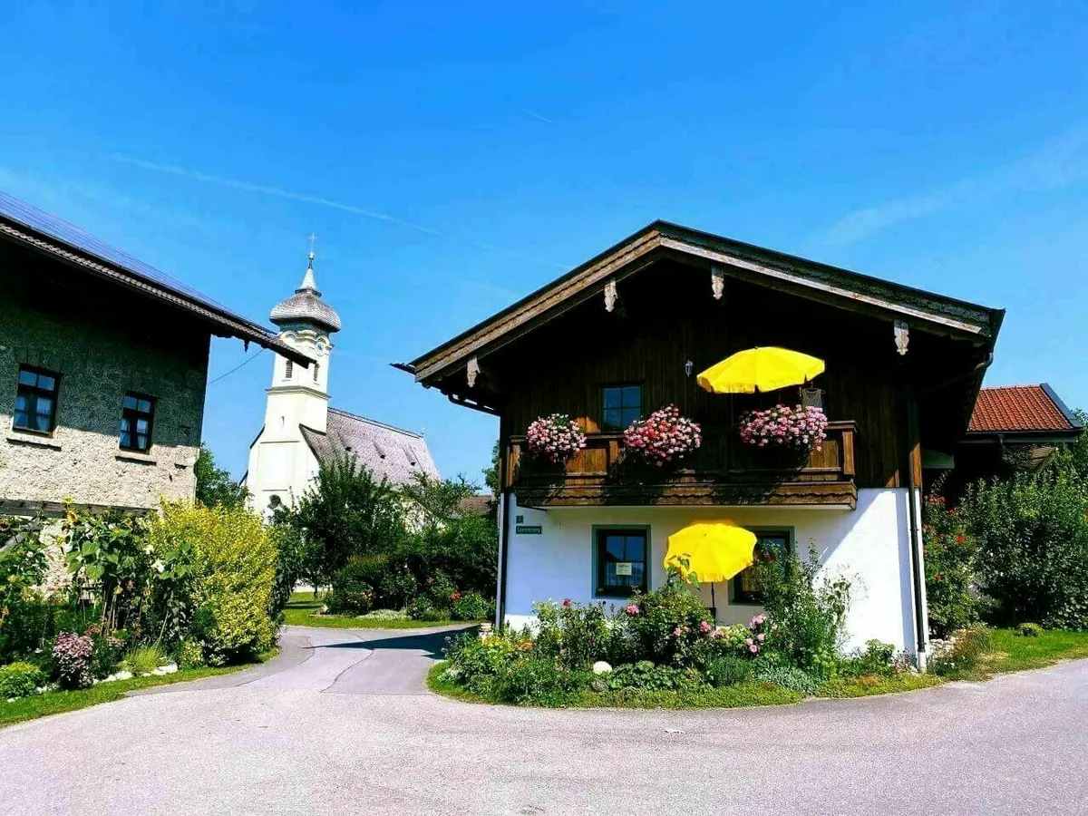 Ein idyllisches, traditionelles bayerisches Haus mit Holzbalkon und Blumenkästen, davor zwei leuchtend gelbe Sonnenschirme. Im Hintergrund ist ein weißer Kirchturm mit Zwiebelhaube zu sehen.