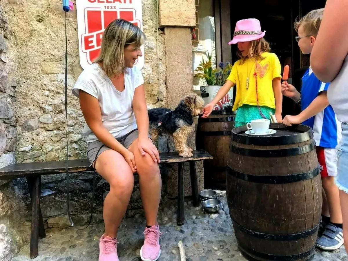 Eine Frau sitzt auf einer Bank im Freien und streichelt einen Hund. Neben ihr stehen zwei Fässer, auf denen eine Tasse und eine Untertasse stehen. Zwei Kinder sind ebenfalls zu sehen, eines hält ein Eis in der Hand.
